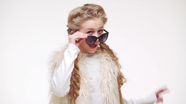 Cool young beautiful Caucasian girl with long blonde hair standing on white background in black sunglasses and confidently smiling in slowmotion