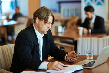 Busy handsome entrepreneur sitting on comfortable armchair in spacious cafe and calling to his business partner, waist-up portrait