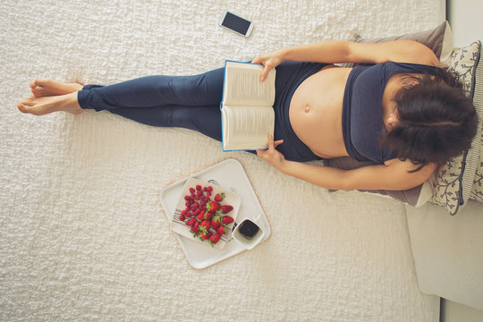 Young pregnant woman, lying in bed with smartphone, book, coffee and fruits,