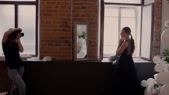 The girl takes off beautiful models in a black wedding dress against the red brick wall near the window. Photo Session behind the scenes.