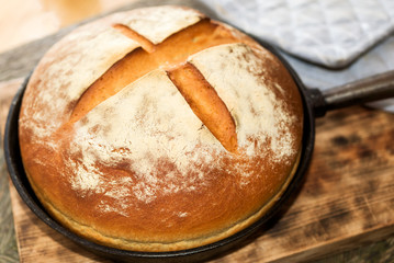 Newly baked artisan bread in cast iron skillet on wooden cutting board. Gray potholder at side.