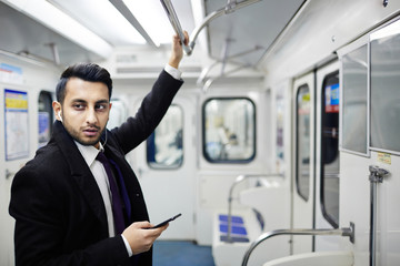 Portrait of handsome Middle-Eastern businessman standing in subway train holding onto hand rail and listening to music from smartphone