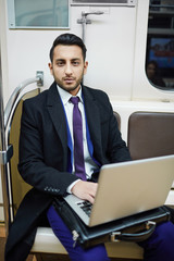 Portrait of handsome Middle-Eastern businessman in subway train, working with laptop and looking at camera