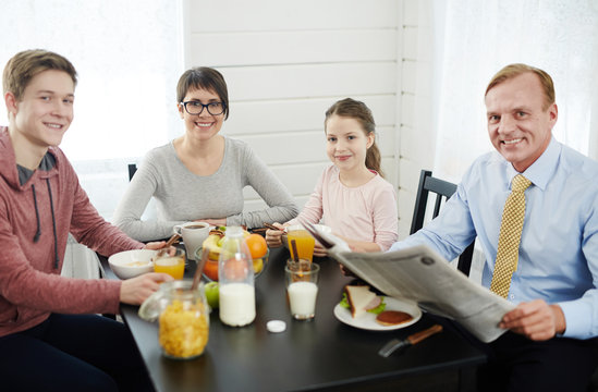 Lovely family of four looking at camera with wide smiles while having healthy breakfast at home, waist-up portrait