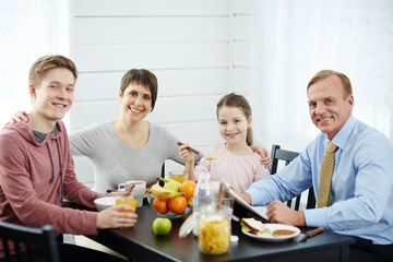 Family portrait in morning kitchen: they looking at camera with toothy smiles, attractive middle-aged mother slightly embracing her children