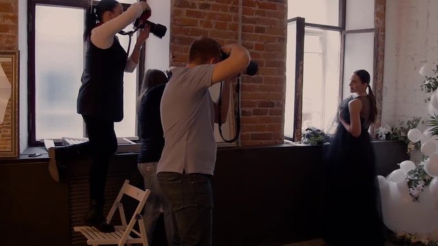 Many photographers on the masterclass take pictures of a beautiful model in a black wedding dress on the background of a red brick wall by the window. Backstage photoshoot.