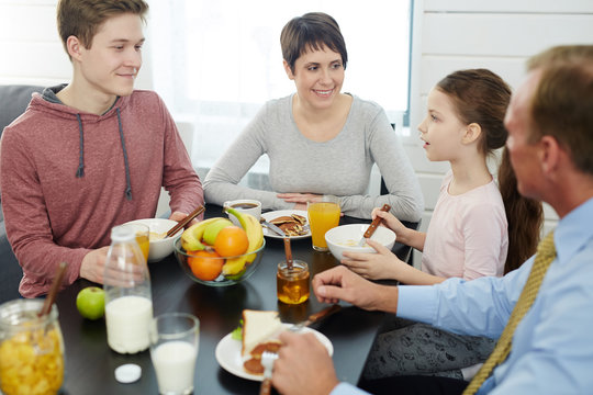 Profile view of cute little girl telling something to her family while eating cornflakes with milk, her father in formalwear, pretty mother and teenage brother listening to her with interest