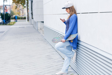Young girl with skateboard leaning on modern gray wall, looking at phone.