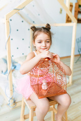 Sweet little girl is hugging a Cage with a bird, looking at camera and smiling while sitting on her сhair at home