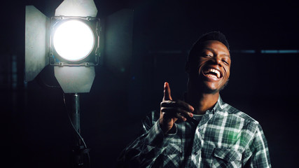 Young African-American man in darkness coming close to camera, then studio light stand is being turned on making man happy and smiling. Looking at camera