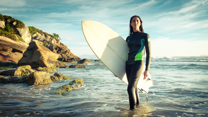 Young female surfer in ocean with surfboard at rocky beach