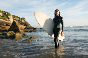 Young female surfer in ocean with surfboard at rocky beach