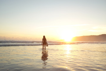 Young female surfer entering the ocean with surfboard at sunset or sunrise