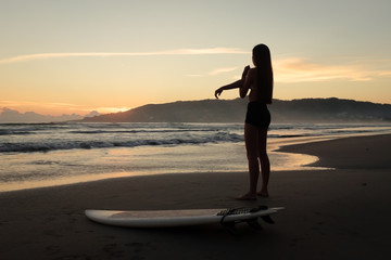 Silhouette of female surfer warming up and stretching at the beach before entering ocean at sunset or sunrise