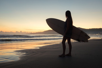 Female surfer with surfboard looking into the ocean during sunset sunrise