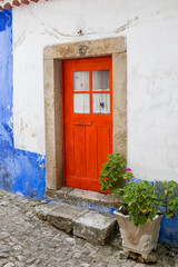 Lovely bright red door with square window, closed white curtains, with a porch of large square stones and a stone vase with a flower.