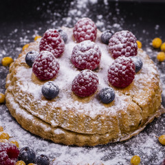 Cake with raspberries, blueberries, sea buckthorn sprinkled powdered sugar on a black plate