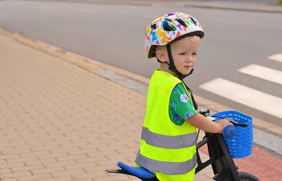 Little boy riding bike in front of zebra crossing. He wears helmet and reflective vest with stripes.