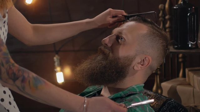 A sexual worker of a barber shop combs a brutal bearded man in Slow motion. The girl in a short dress sits on a brutal man and shaves him with a dangerous razor.