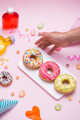 Party. Hand holding plate of colourful sugary round glazed donuts and bottles of drinks on pink background.