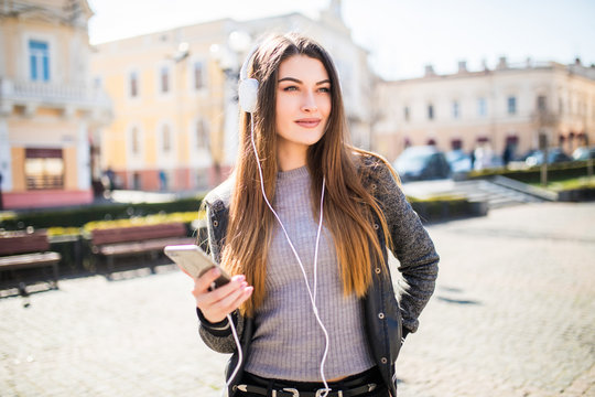 Portrait of happy girl listening music on line with wireless headphones from a smartphone in the street in sunny day