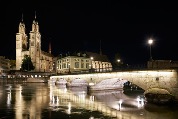 Fototapeta na wymiar Nachtaufnahme Zürich mit beleuchteter Münsterbrücke und dem Grossmünster, mit Wasserspiegelungen in der Limmat