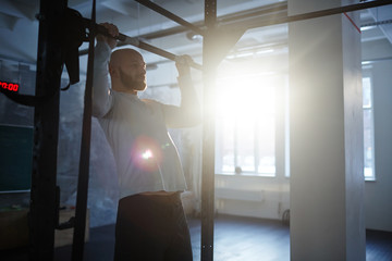 Portrait of strong bearded man performing pull ups on bar during workout in gym lit by sunlight
