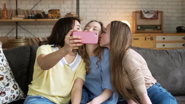 three young female best friend have fun and make selfie, grimacing and laughing using telephone mobile camera, while sitting at home on sofa