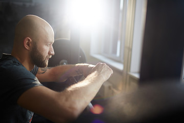 Side view portrait of modern bald bearded man looking out of window pensively in gym, leaning on weights rack in sunlight