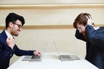 Cheerful entrepreneur sitting at one end of table and holding fist up while his colleague clutching head in despair and sitting at another end