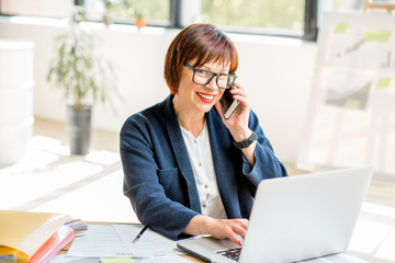 Senior businesswoman working with laptop and talking phone at the bright modern office interior