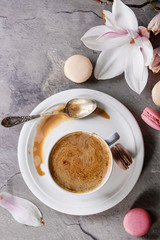 White cup of black coffee, served on white saucer with macaroons biscuits, spoon and magnolia flower blossom branch over gray texture background. Flat lay, space