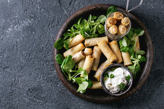 Fried spring rolls with white yogurt sauces, served in terracotta plate and fry basket with fresh green salad over black texture background. Flat lay, space. Asian food