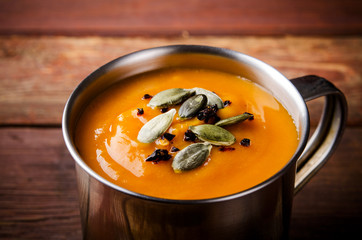 Roasted pumpkin soup in metal mug with seeds and smoked paprika, on rustic wooden background.