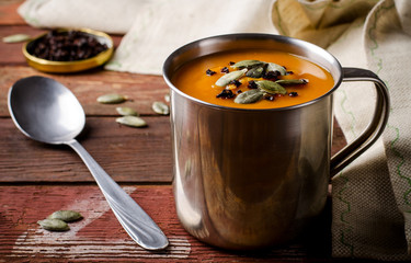 Roasted pumpkin soup in metal mug with seeds and smoked paprika, on rustic wooden background.