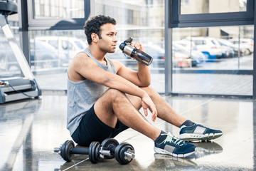Fototapeta na wymiar Handsome young man with bottle of water resting on floor at gym