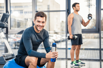 Fototapeta na wymiar Handsome young man with bottle of water resting at gym, other man workout on blurred background