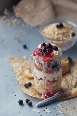Layered dessert with muesli, yogurt, banana, linen seeds, sliced almonds and berries in glass mason jar on blue rustic background.