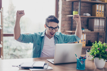 Excited screaming young man looking at the screen of his computer and triumphing with raised hands