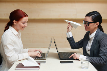 Two playful pretty financial managers sitting opposite one another, preparing annual accounts on laptops and launching paper planes