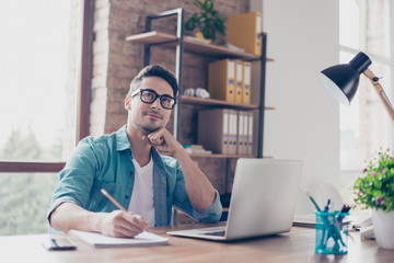 Portrait of minded young student sitting at the table in front of computer and writing a...