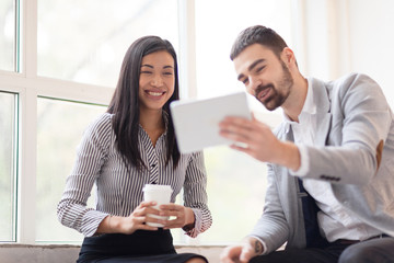 Spending Coffee Break with Colleague: cheerful Asian employee holding paper cup of coffee in hands and sitting on windowsill, her handsome colleague showing her something on digital tablet