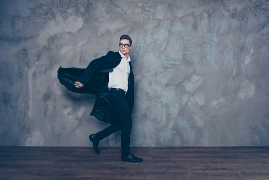 Young attractive man in glasses and black coat is rushing somewhere and looking back. He is in formal wear, behind is grey background