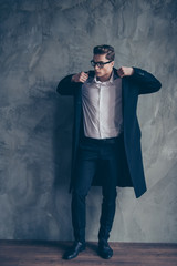Attractive businessman in glasses is fixing his black coat. He is standing near the grey wall. Looks confident and successful