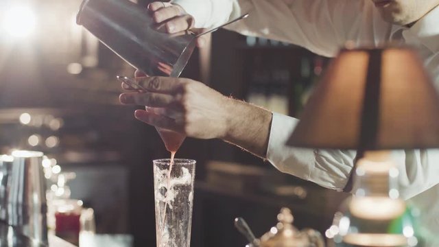 20s Bartender in a hat is pouring mixed cocktail from shaker in a glass