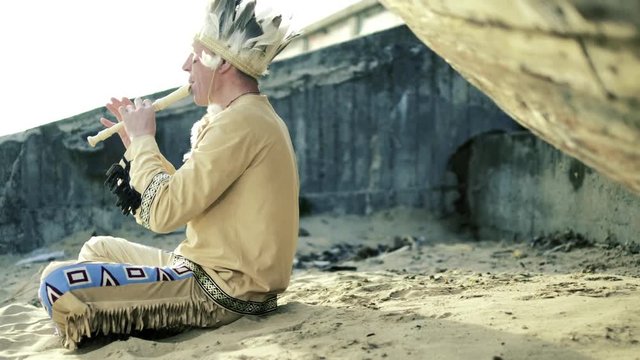 An Indian in a national costume plays a flute near a boat on the river bank 4k