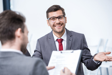 Smiling man in suit and eyeglasses gesturing and looking at businessman with clipboard, business concept