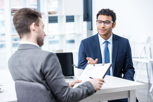 Smiling man in suit and eyeglasses gesturing and looking at businessman with clipboard, business concept