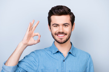 Cheerful young handsome man in jeans shirt on light blue background is showing "OK" gesture, smiling