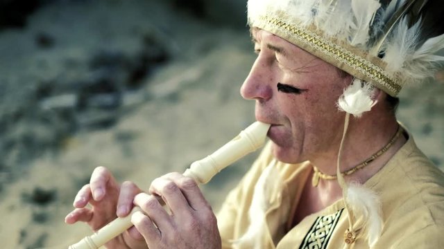 An Indian in a national costume plays a flute near a boat on the river bank 4k